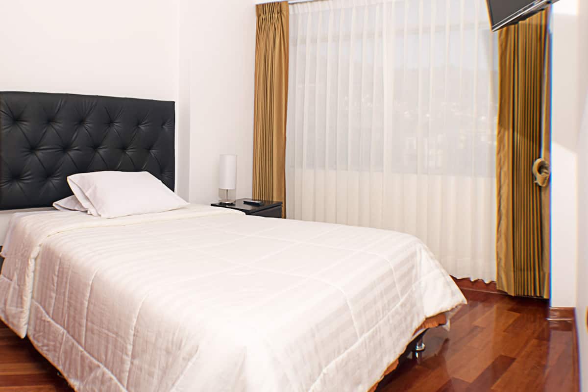 A neatly made bed with a black headboard, a bedside table with a lamp, and a window with sheer curtains.