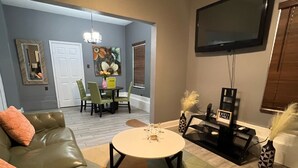 A living room with a green sofa, a coffee table, a TV mounted on the wall, and a dining area in the background. - No Place Like a Home Away From Home (New Orleans)