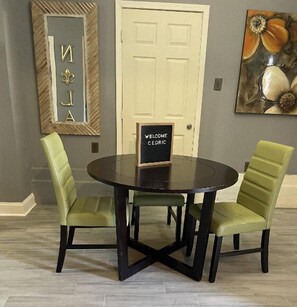 Een eetgedeelte met een ronde tafel en twee stoelen, een welkomstbord, een deur en een spiegel met decoratieve letters.