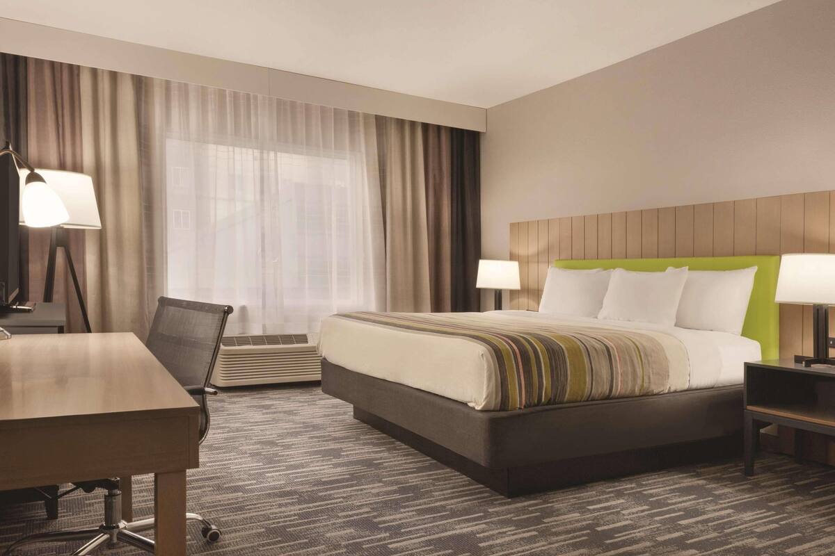 A hotel room with a large bed, a desk, a chair, a lamp, and a window with curtains.