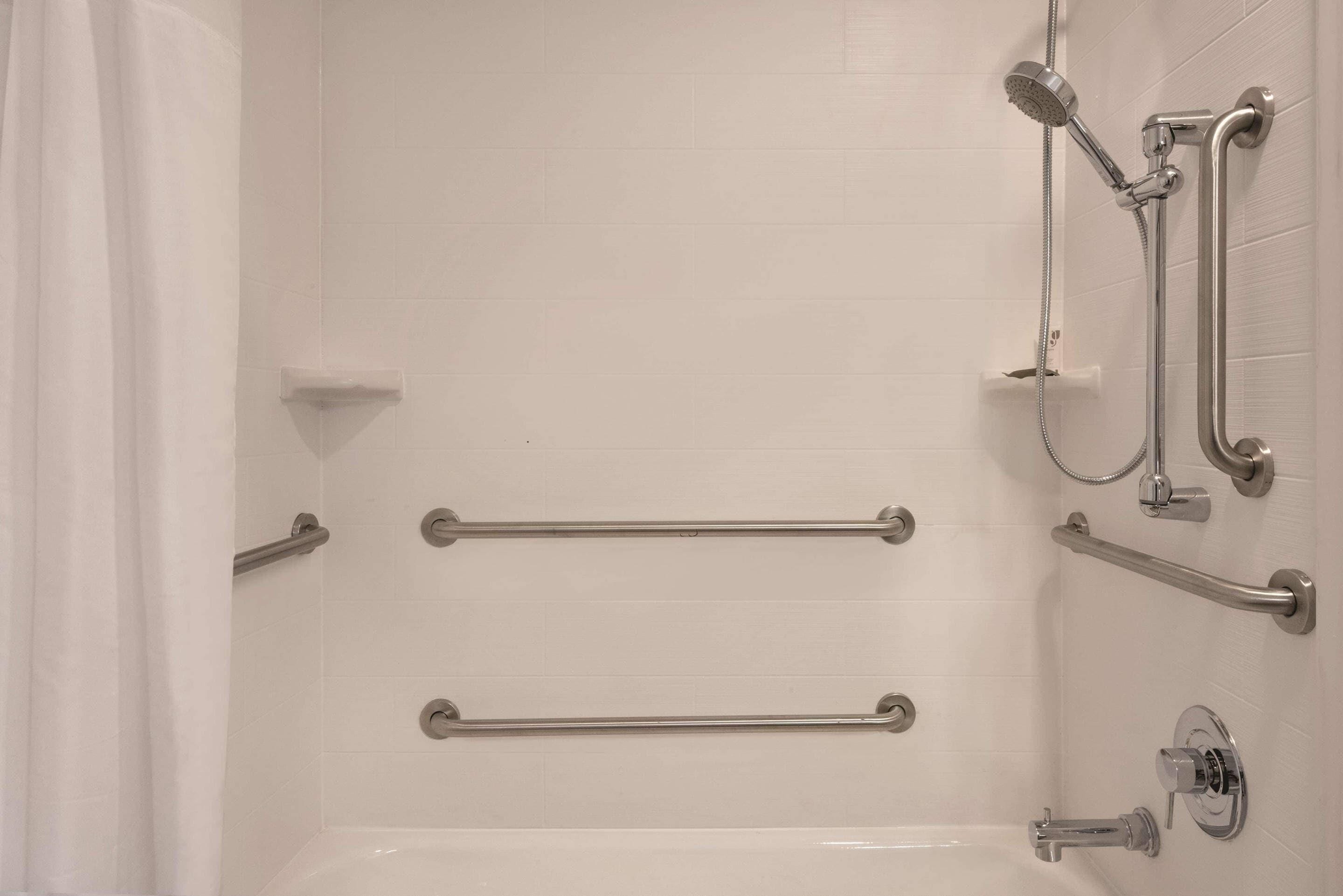 A white tiled shower area with grab bars and a handheld showerhead.