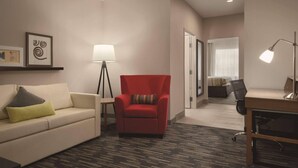 Une chambre d’hôtel avec un fauteuil rouge, un canapé blanc avec des coussins, un bureau avec une lampe et une petite table également dotée d’une lampe.