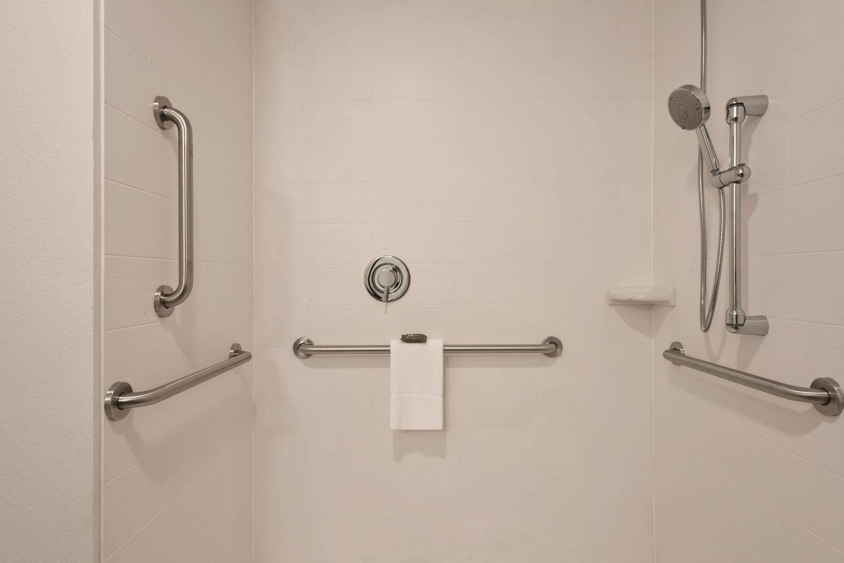 A bathroom with white tiled walls, a white towel hanging on a metal rack, and a shower with a handheld showerhead.