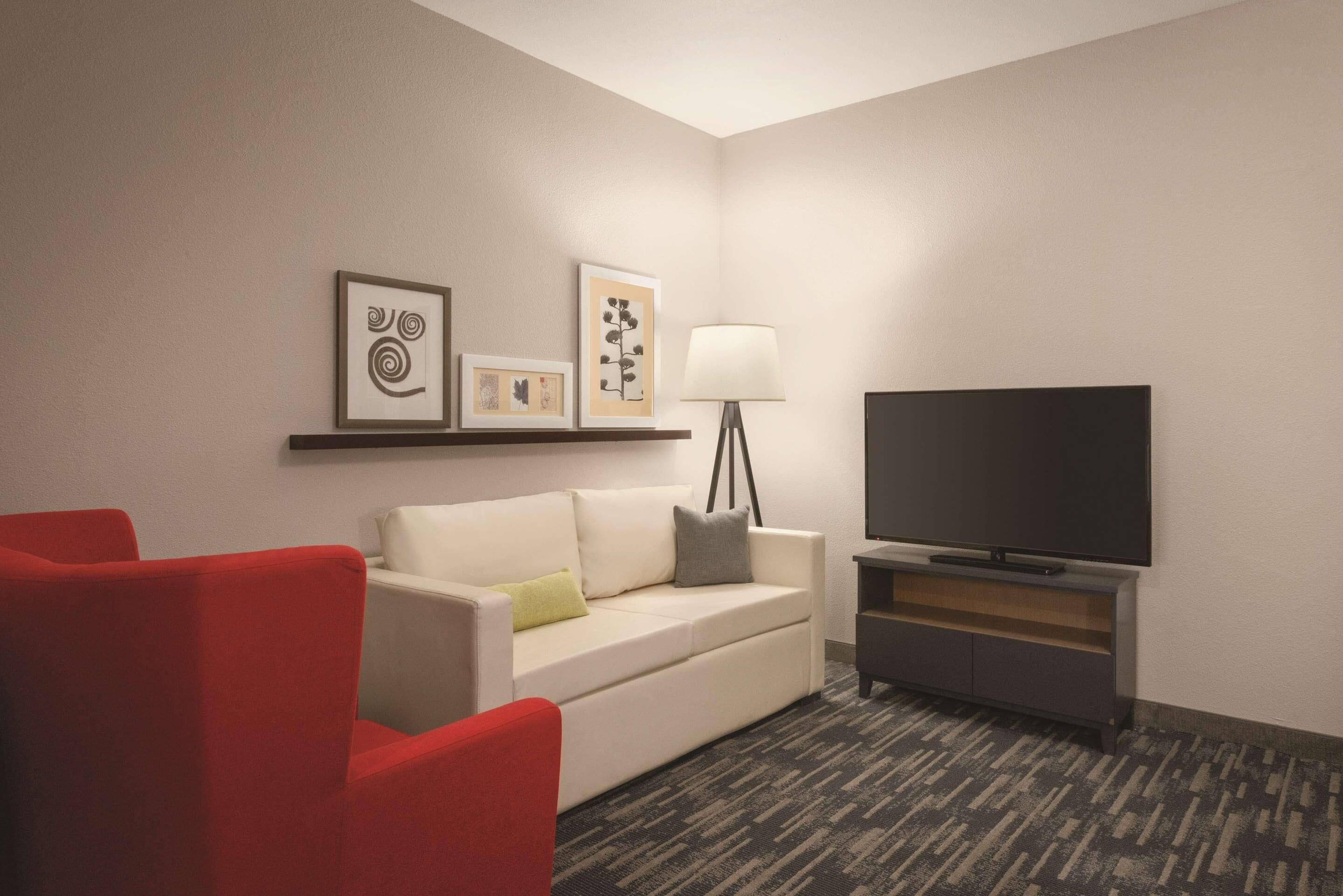 Habitación de hotel con sofá blanco, sillón rojo y televisor de pantalla plana sobre un soporte.