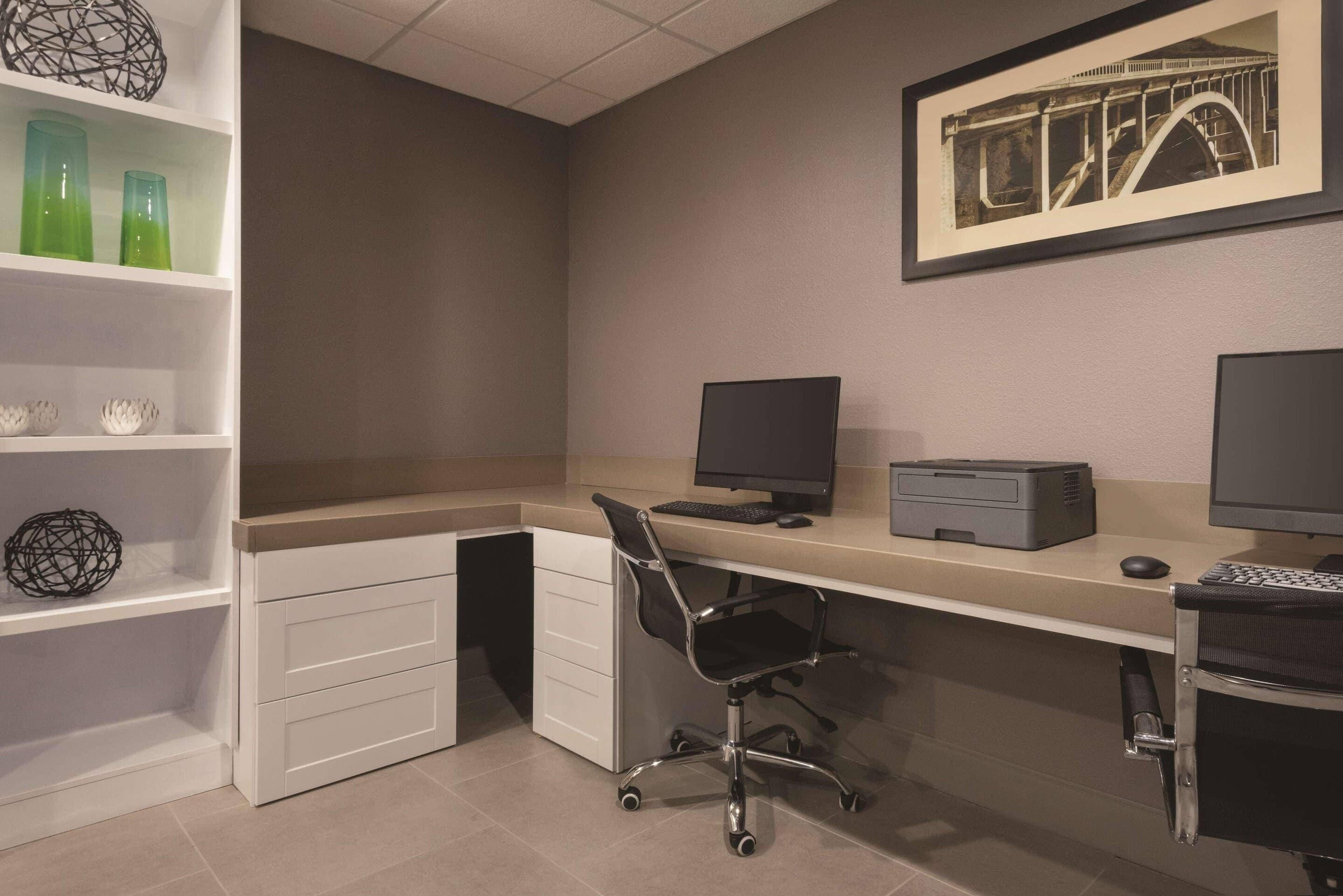 A modern office space with a desk, computer, printer, and a chair. There are shelves with decorative items and a framed picture on the wall.