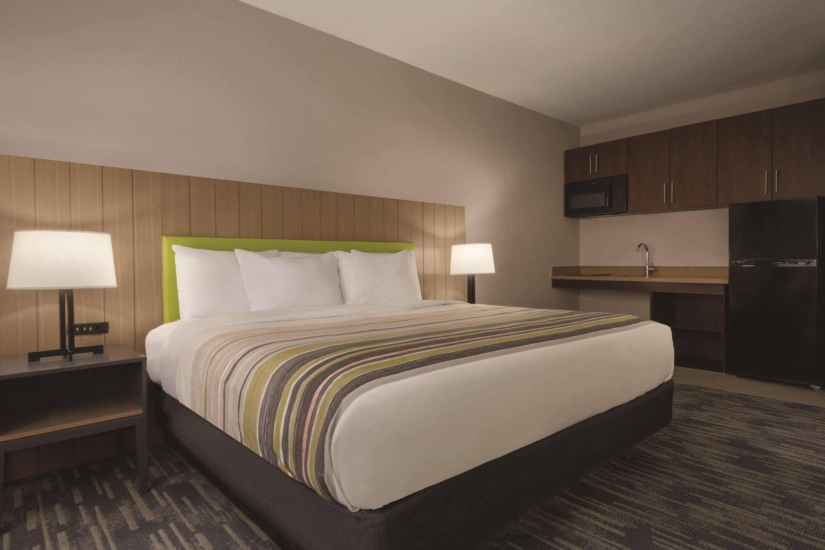 A hotel room with a large bed, two bedside tables with lamps, a wooden headboard, a kitchenette with a microwave and refrigerator, and a carpeted floor.