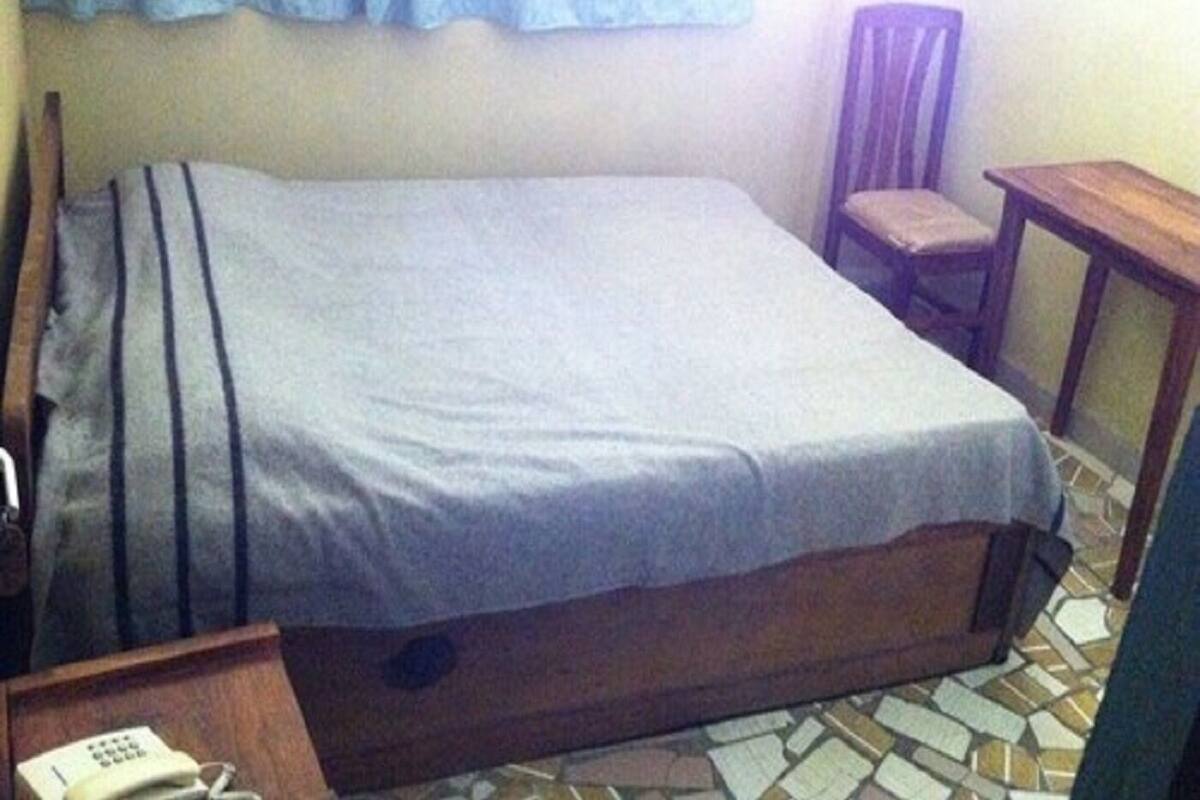 Una habitación pequeña con una cama individual, estructura de madera, una silla, una mesita y un teléfono.