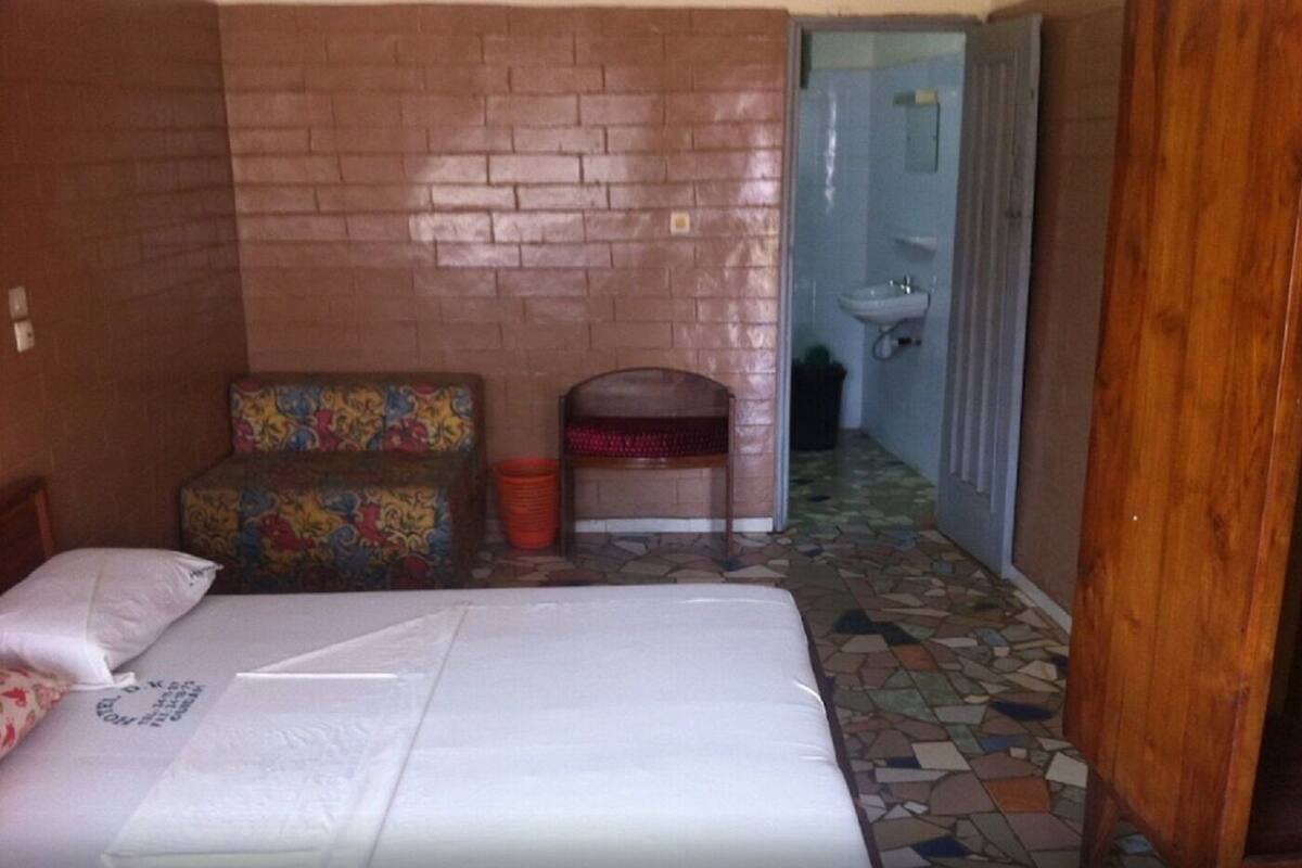 Una habitación con una cama, un sofá colorido, un pequeño banco, un cubo de basura y un baño visible a través de una puerta abierta.