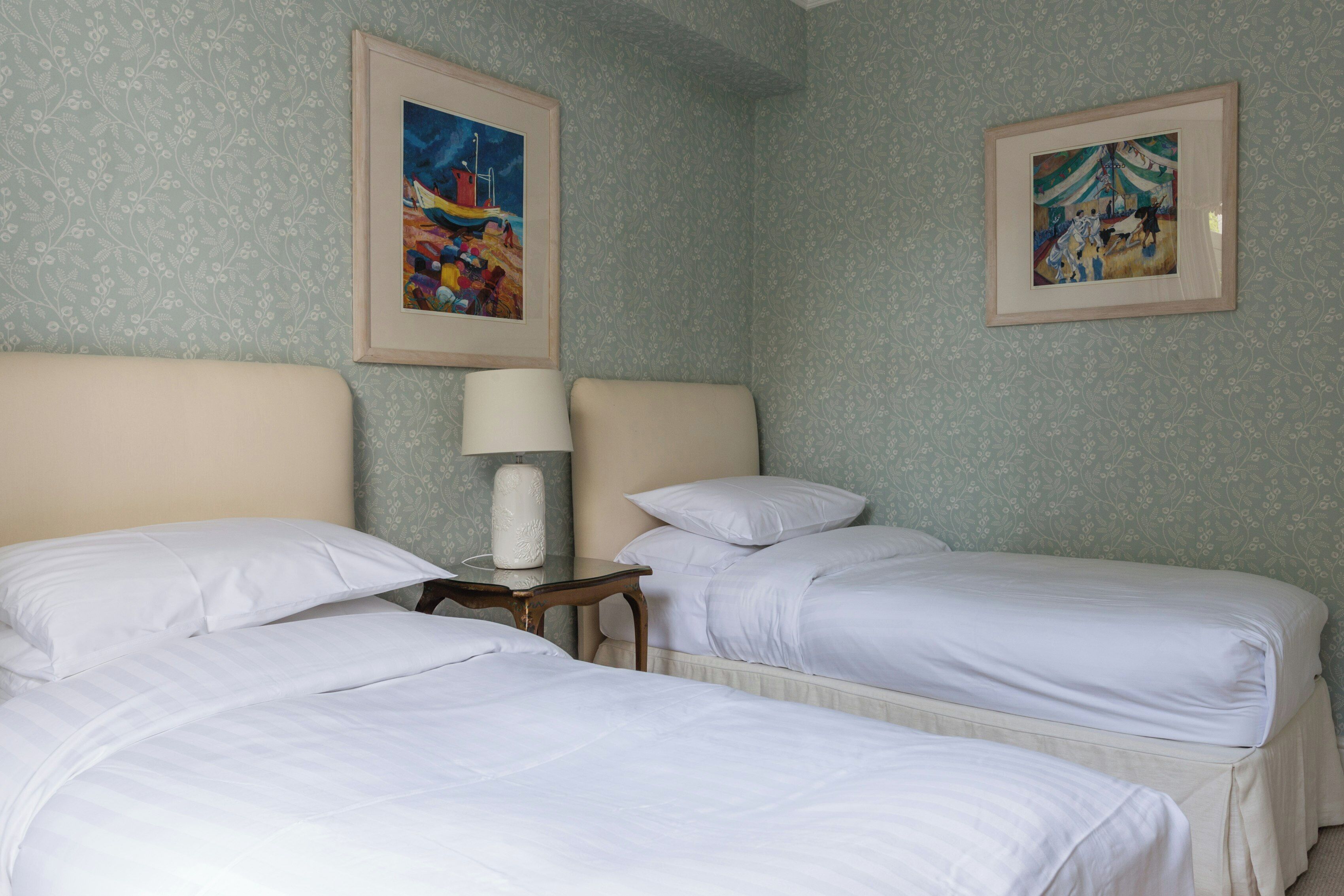 A hotel room with two beds, a nightstand, a lamp, and two framed pictures on the wall.