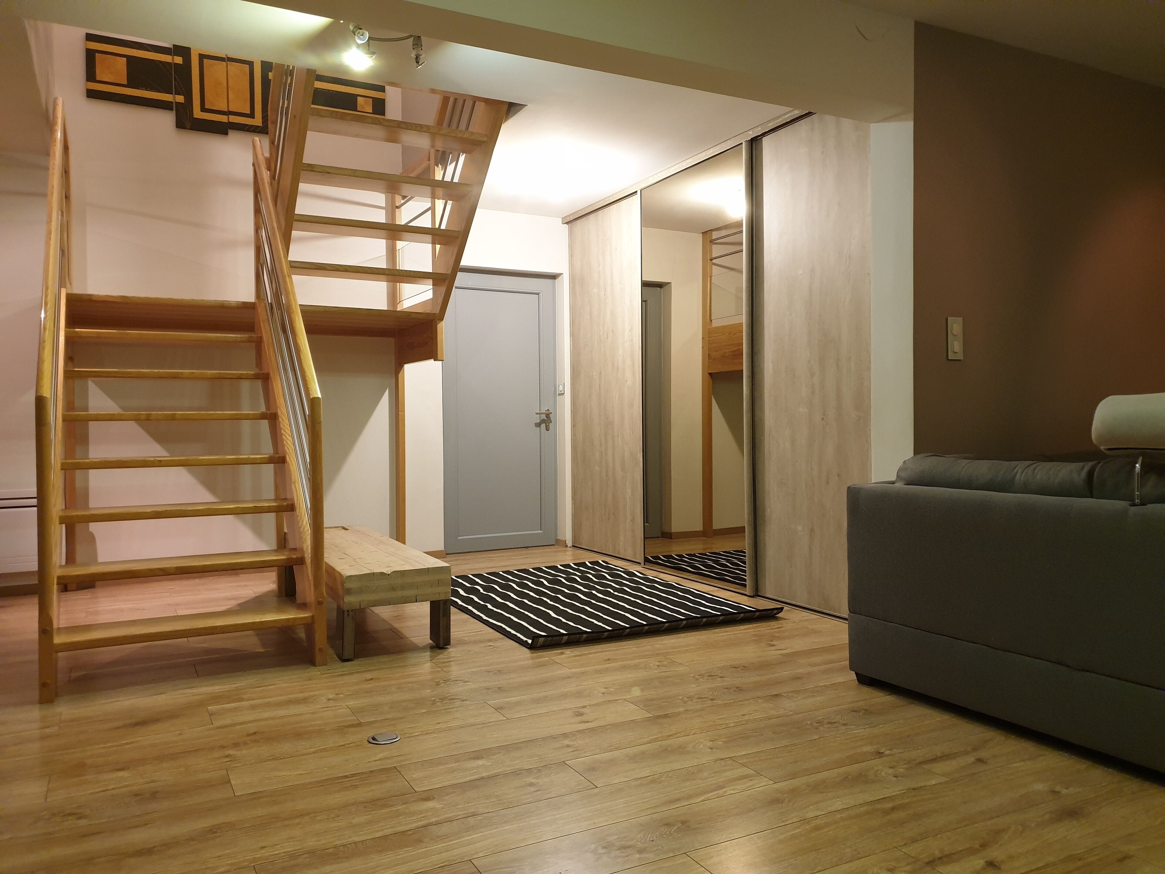 Une pièce avec un escalier en bois, un canapé gris et une porte qui donne sur une autre pièce.