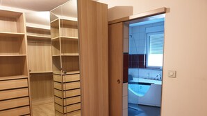 Une chambre moderne avec une armoire en bois, un placard avec des portes miroir et une vue sur une salle de bain équipée d’une baignoire et d’un lavabo.