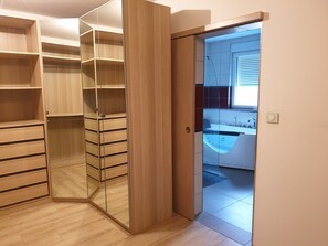 Une chambre moderne avec une armoire en bois, un placard avec des portes miroir et une vue sur une salle de bain équipée d’une baignoire et d’un lavabo.