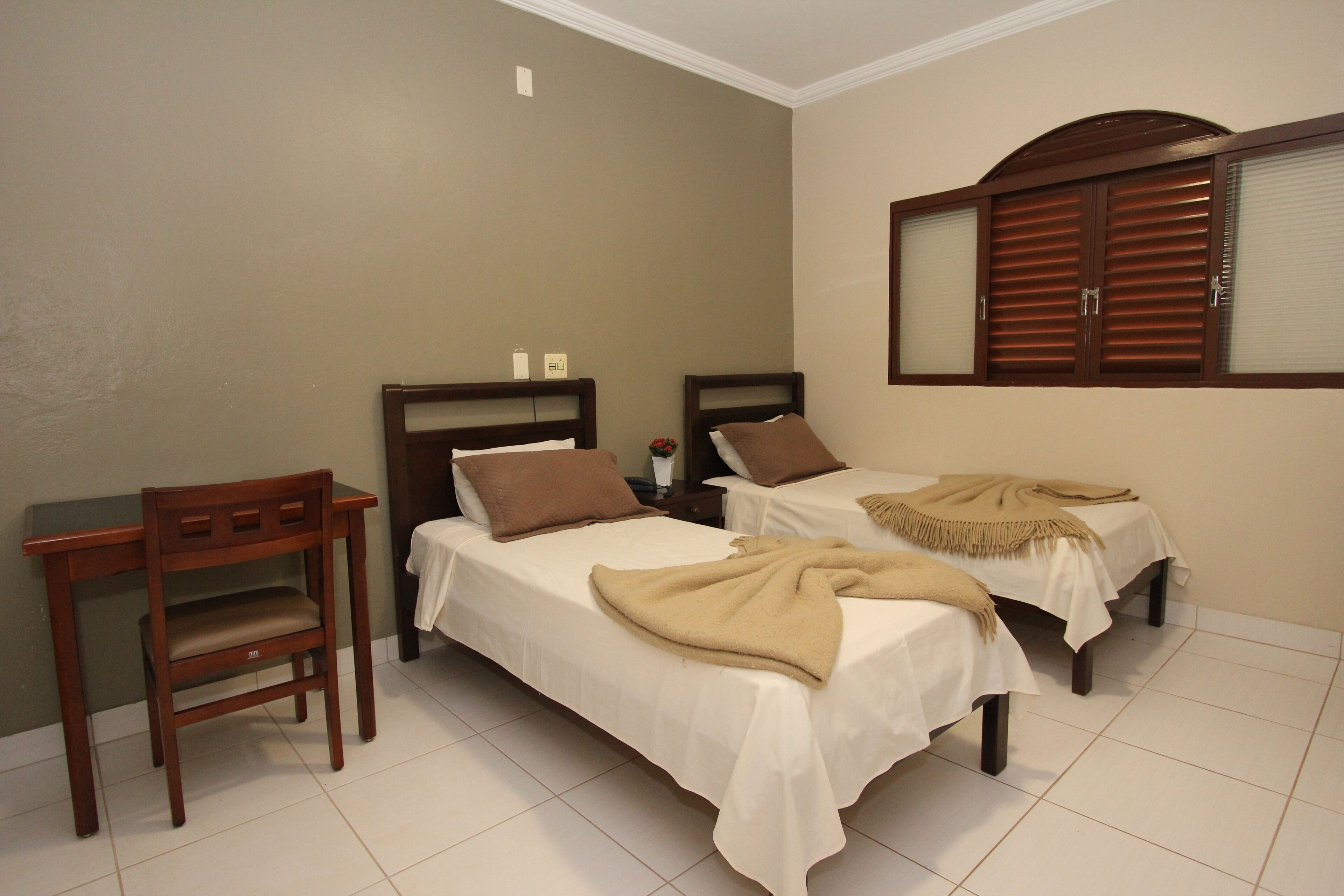Quarto com duas camas de solteiro, uma escrivaninha de madeira e um espelho com venezianas de madeira.