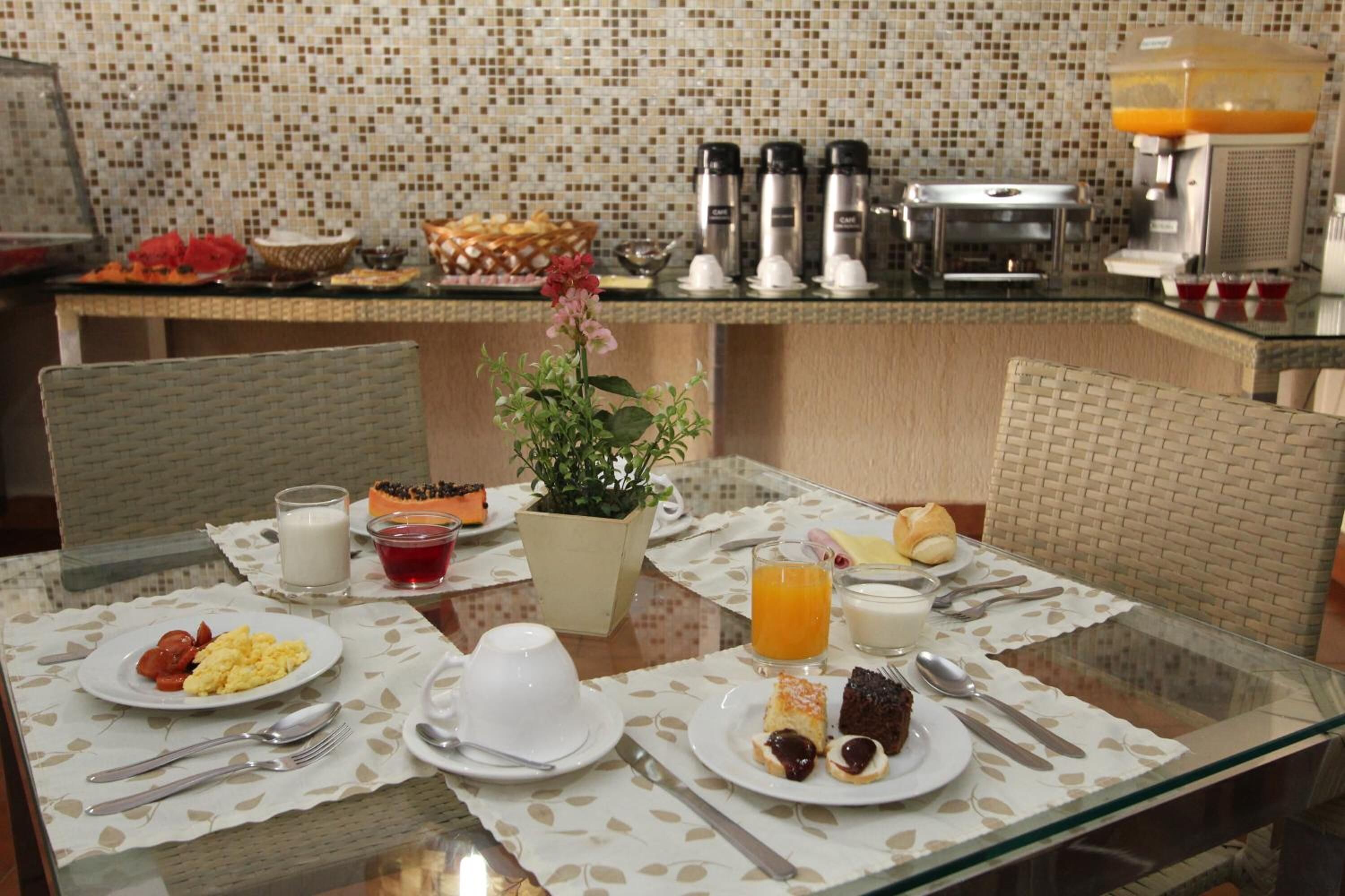 Une table en verre dressée pour le petit-déjeuner, avec des assiettes remplies de nourriture, un arrangement floral au centre et une machine à café en arrière-plan.