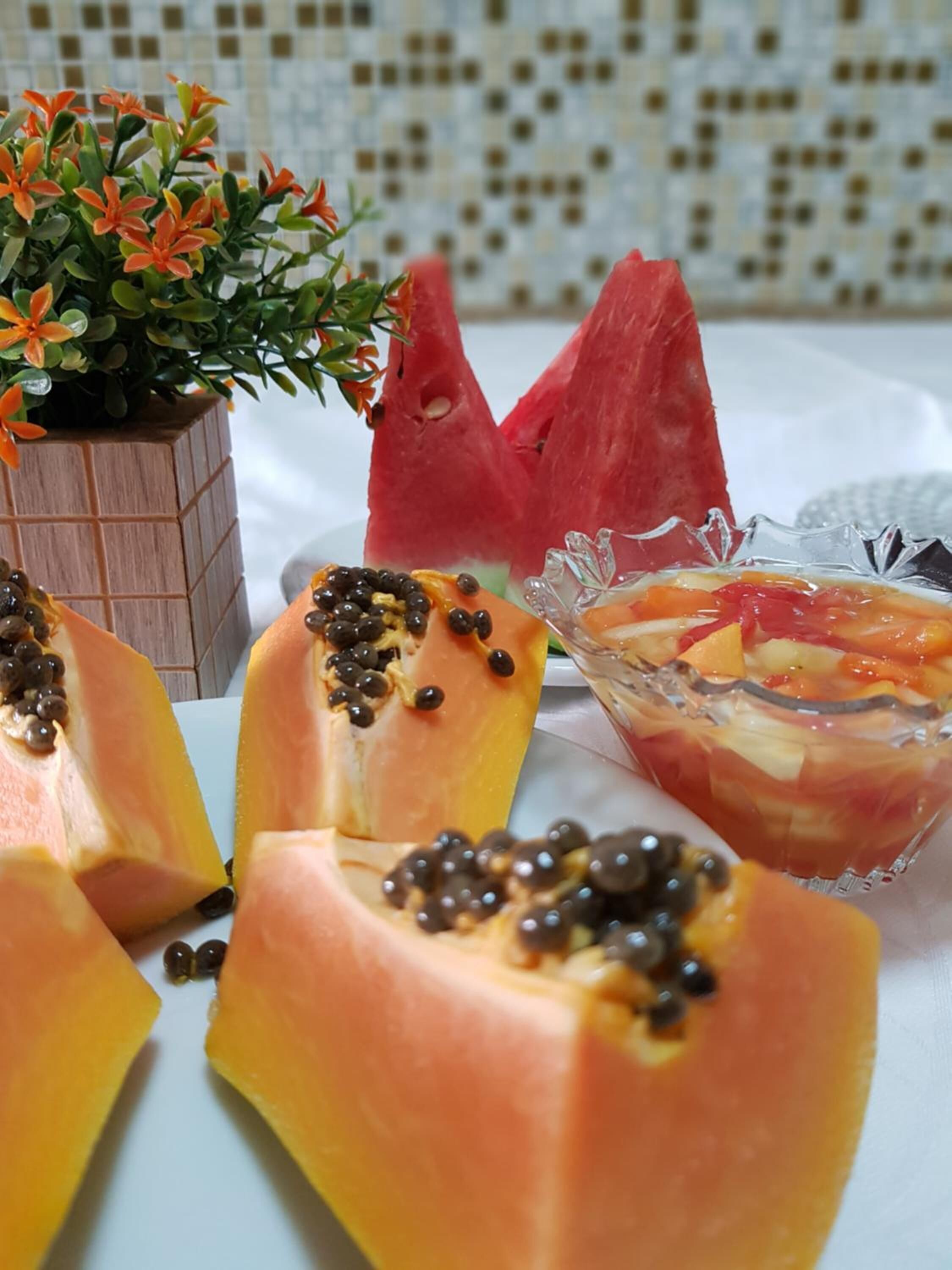 A table with a glass bowl containing a fruit salad, a carved watermelon, and sliced papaya with black seeds.
