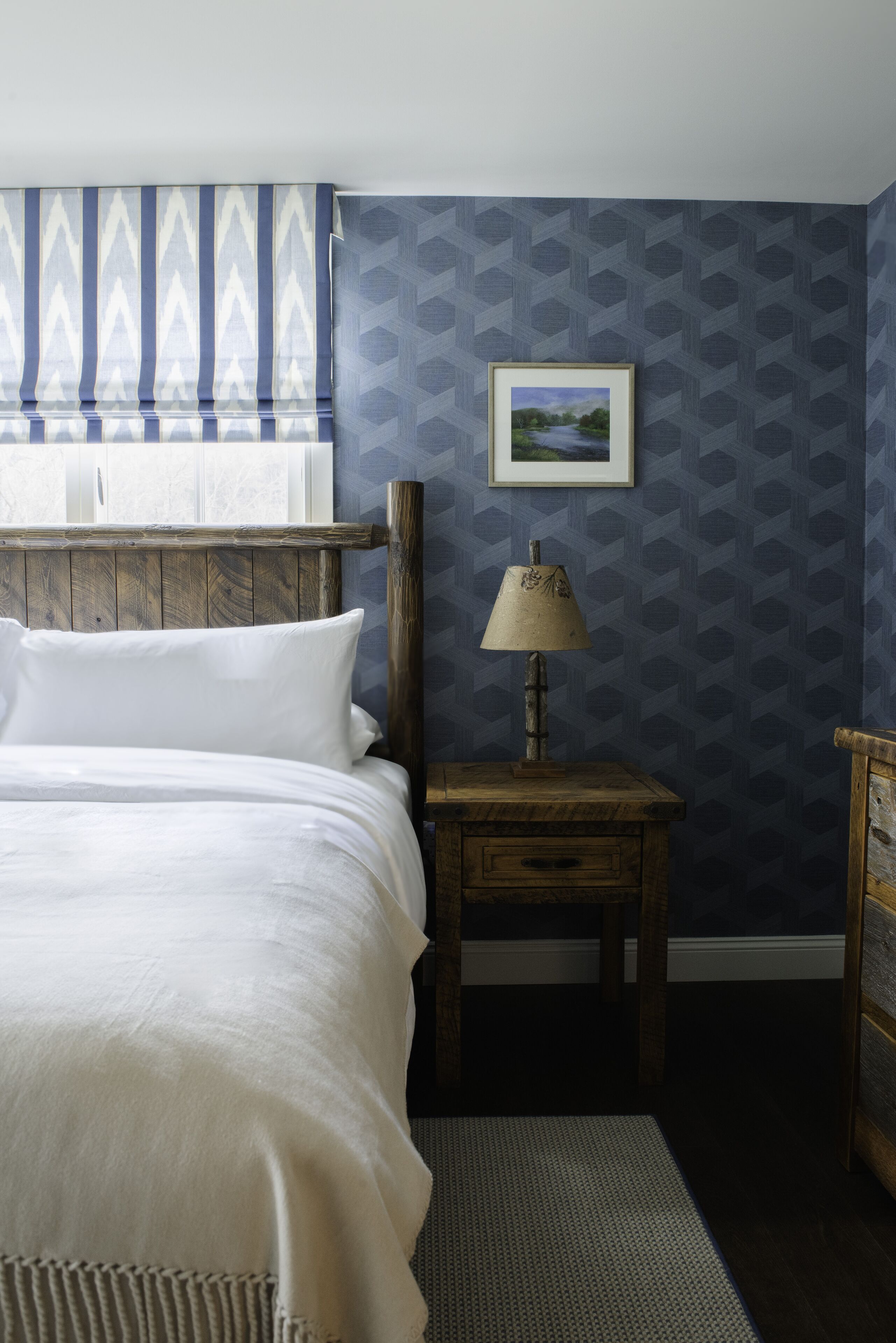 A bedroom with a wooden headboard, a bed with white linens, a nightstand with a lamp, and a framed picture on the wall.