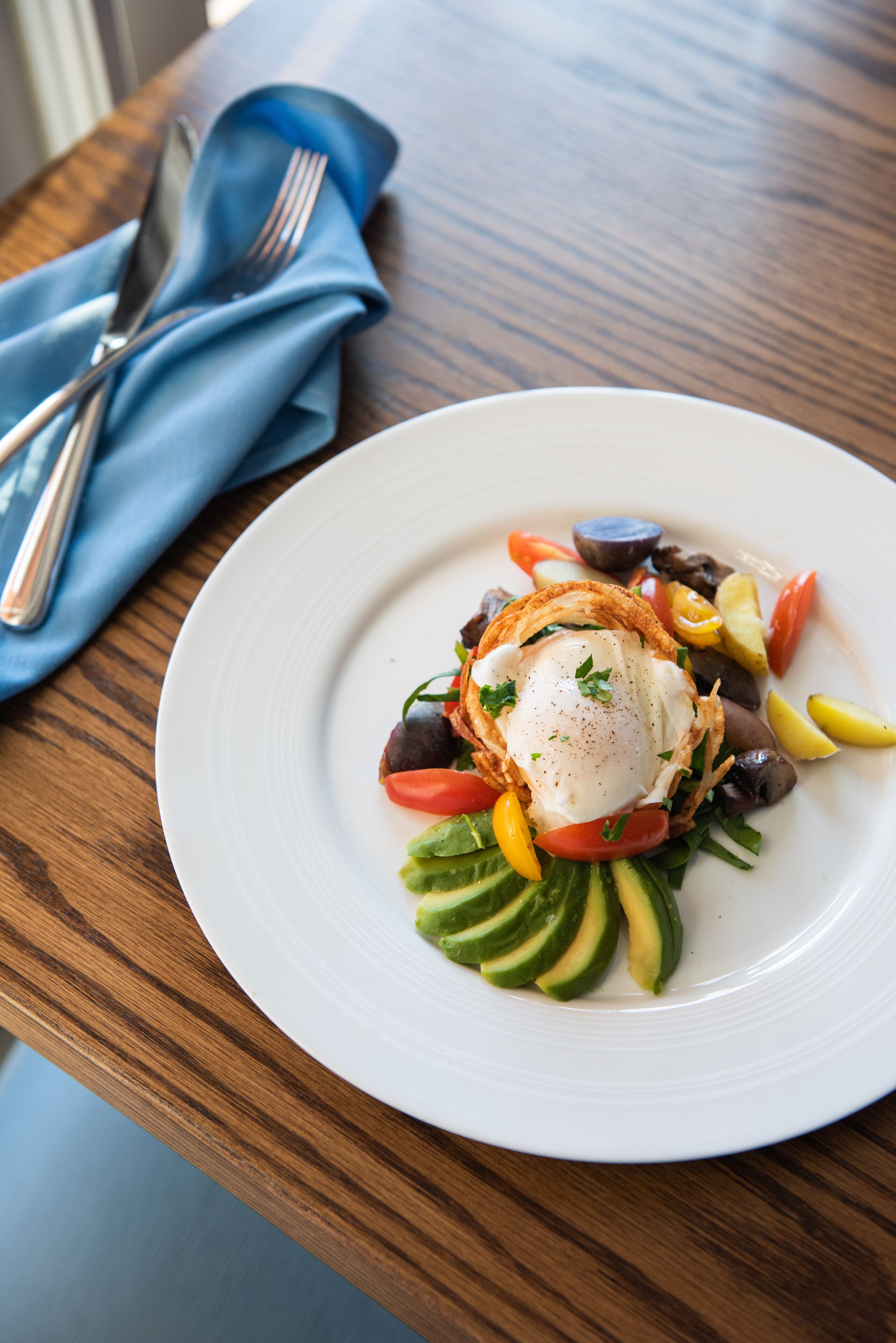A white plate with a poached egg, avocado slices, and mixed vegetables on a wooden table.