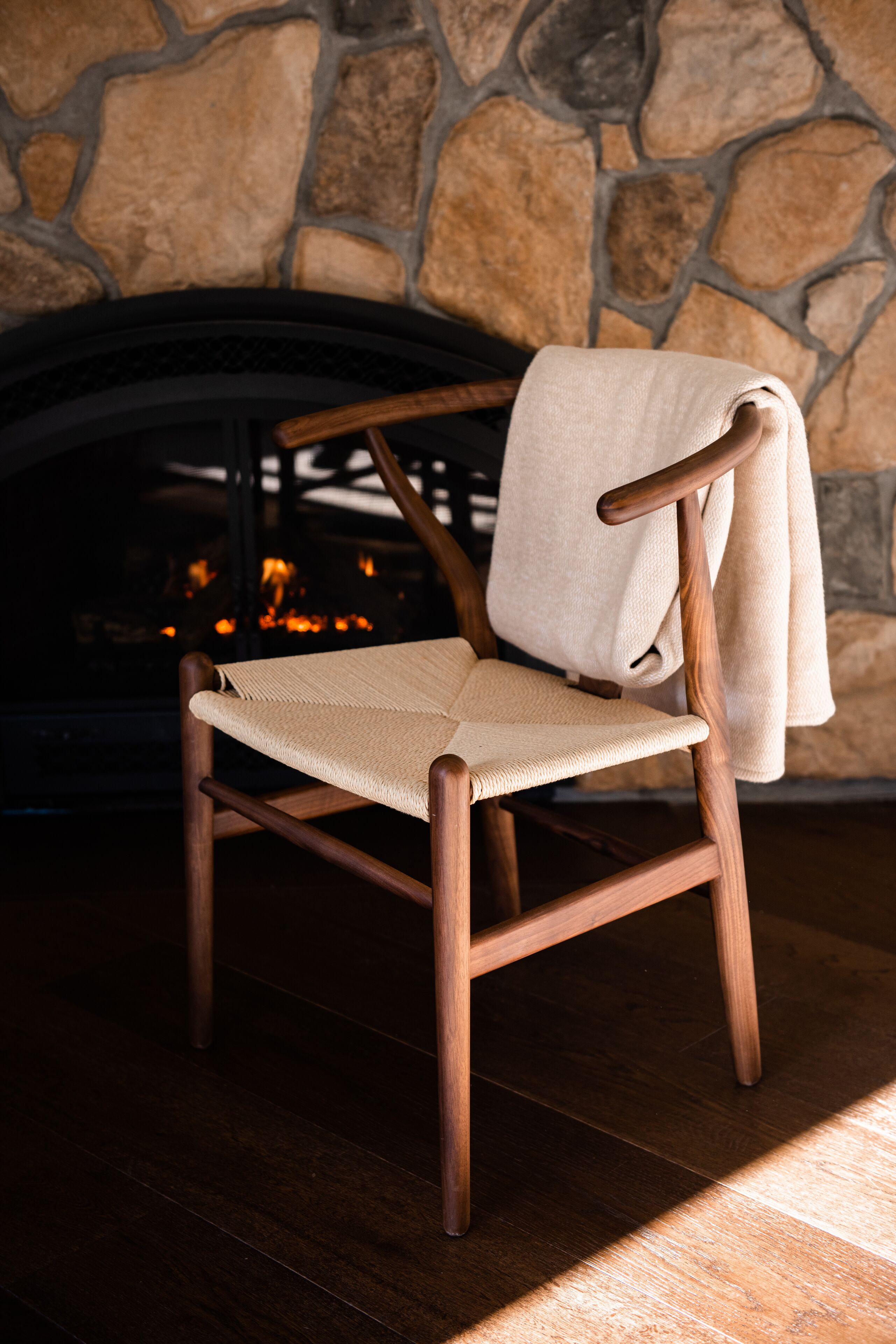 A wooden chair with a woven seat and backrest, positioned in front of a stone fireplace with a visible fire.