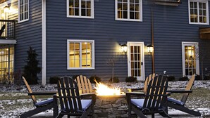 Kaksikerroksinen talo, jossa on kivinen takka, kaksi Adirondack-tyylistä puutarhatuolia ja lumipeitteinen nurmikko.