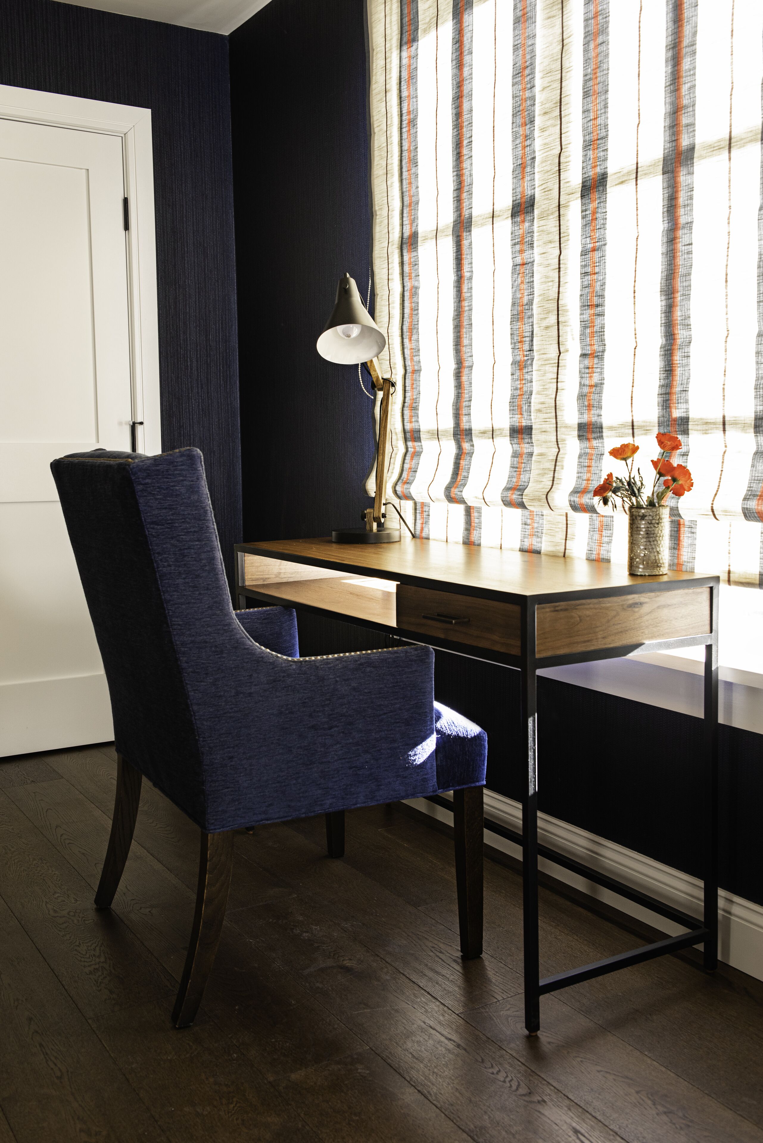 A room with a wooden desk, a blue chair, a lamp, and a window with striped curtains.