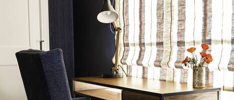 A room with a wooden desk, a blue chair, a lamp, and a window with striped curtains.