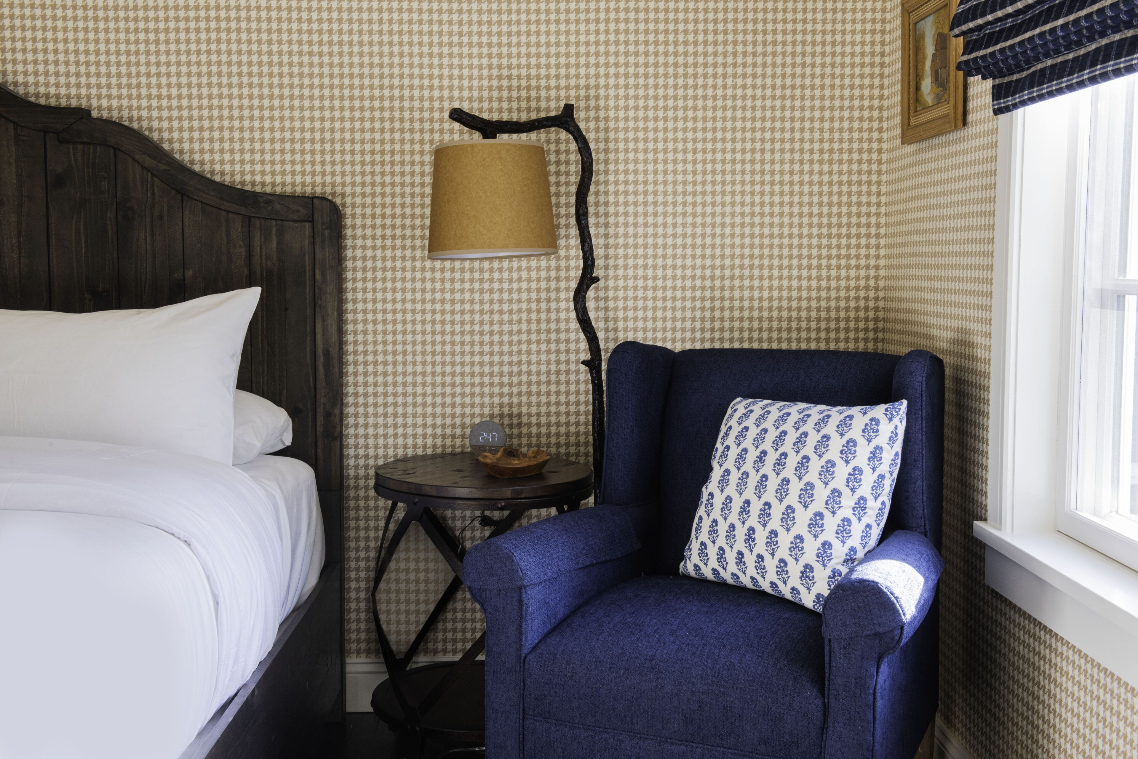 A bedroom with a bed, a blue armchair with a patterned pillow, a small table with a lamp, and a window with curtains.