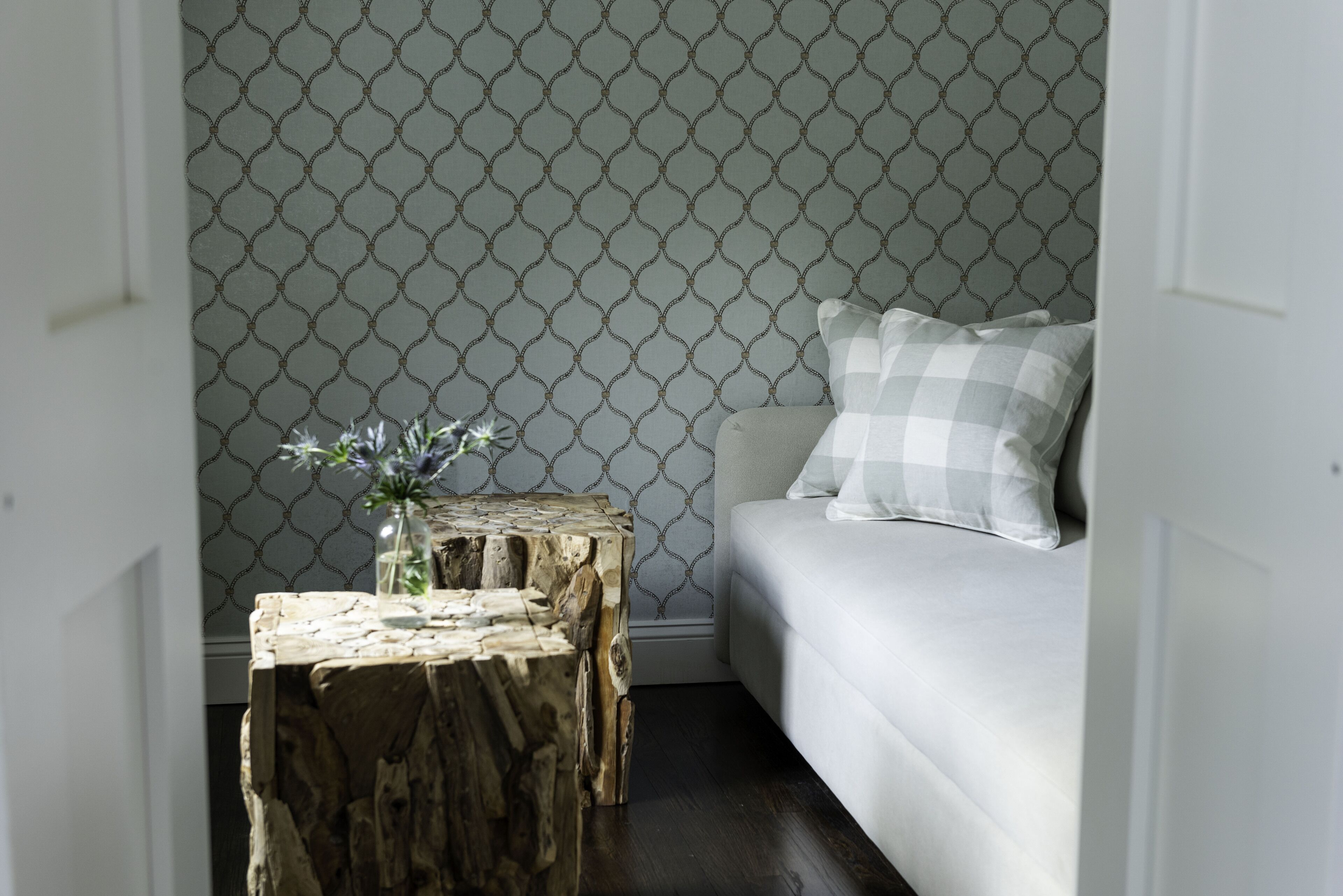 A room with a patterned wallpaper, a wooden side table with a vase, and a white sofa with a checkered pillow.