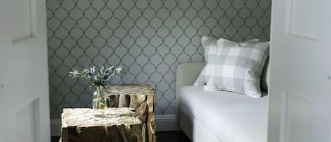 A room with a patterned wallpaper, a wooden side table with a vase, and a white sofa with a checkered pillow.