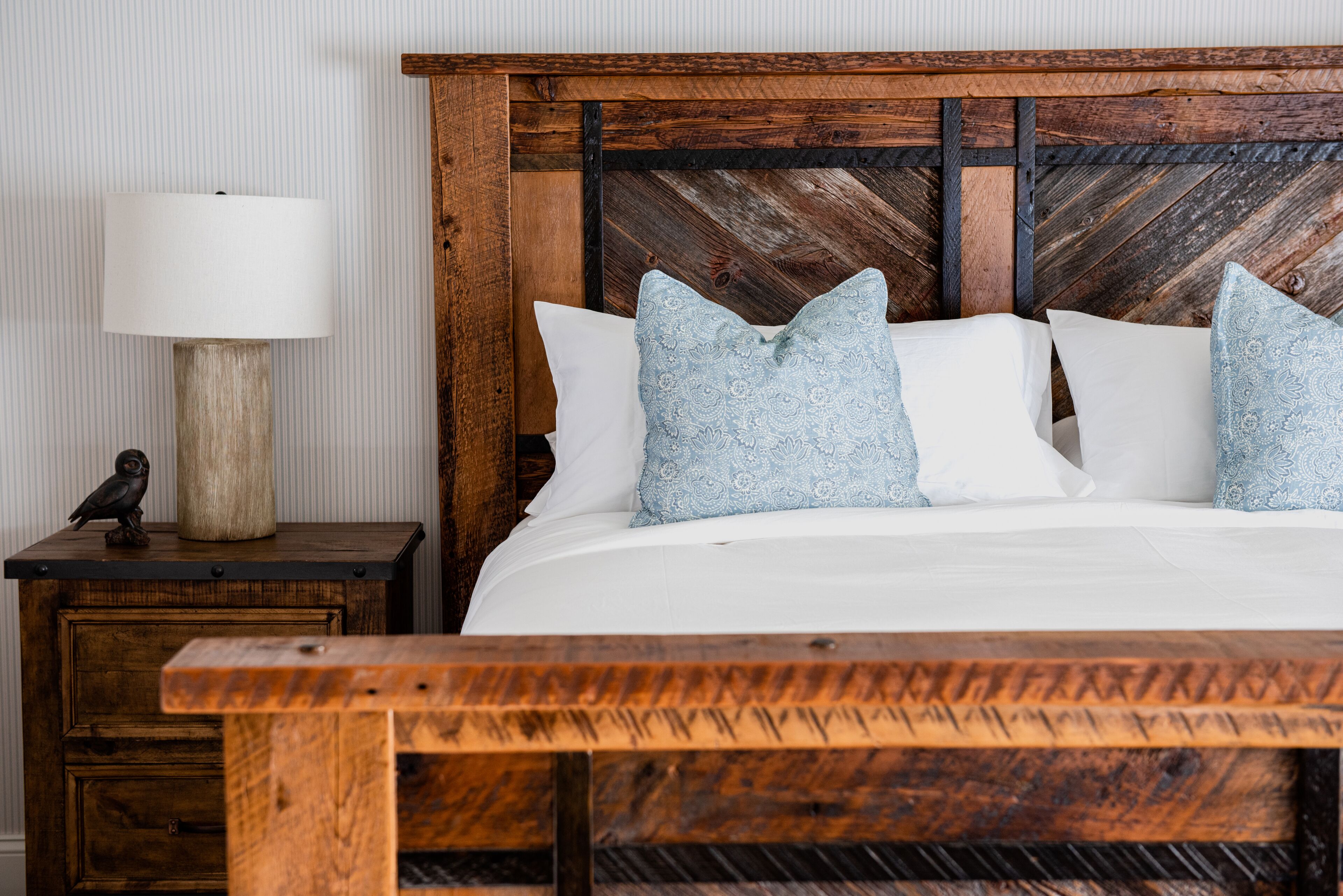 A wooden bed with white bedding and light blue pillows, a wooden nightstand with a lamp, and a small wooden table with a bird figurine.