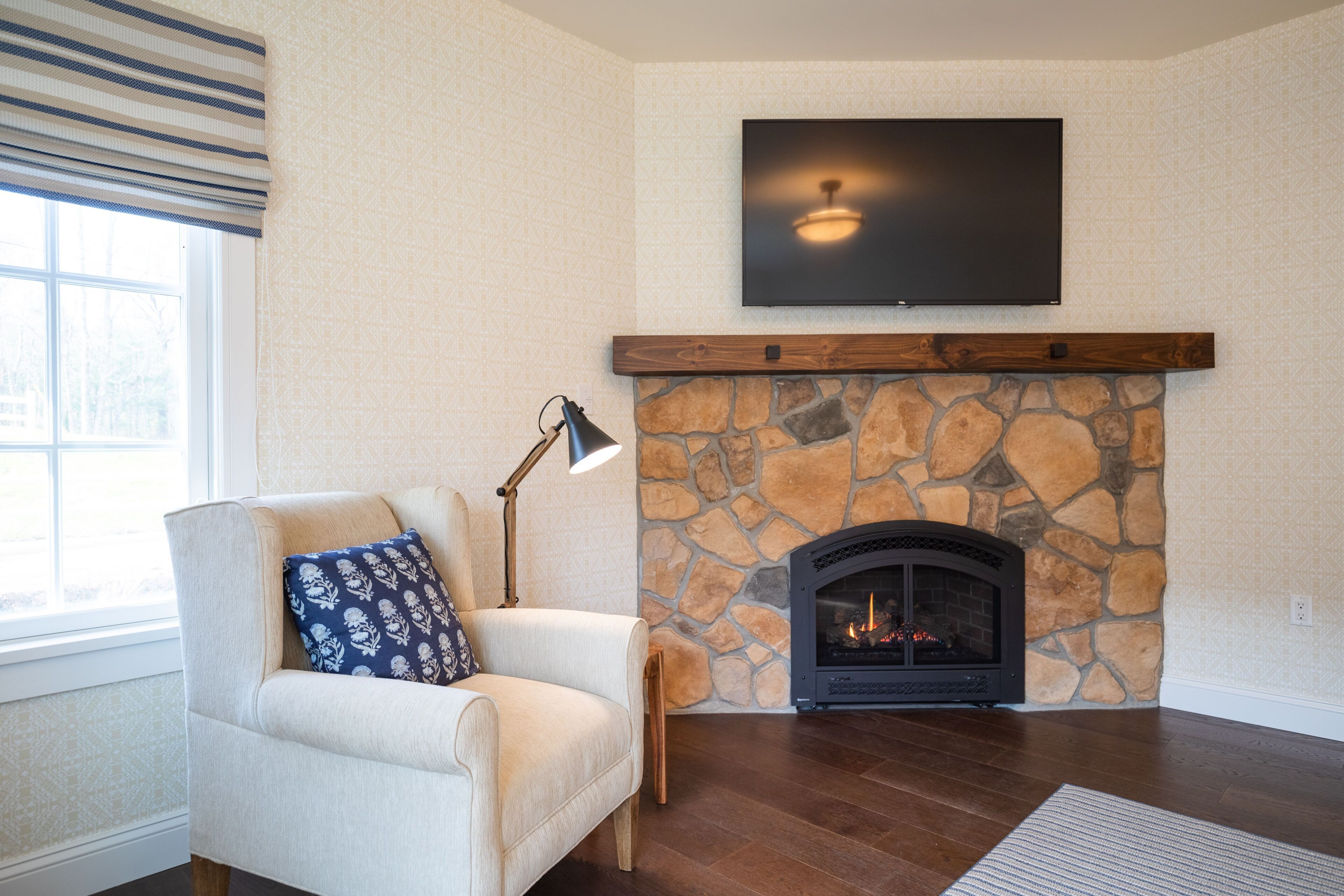 A living room with a stone fireplace, a sofa with a patterned pillow, a floor lamp, and a flat-screen TV mounted above the fireplace.