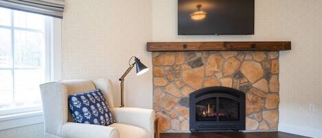 A living room with a stone fireplace, a sofa with a patterned pillow, a floor lamp, and a flat-screen TV mounted above the fireplace.