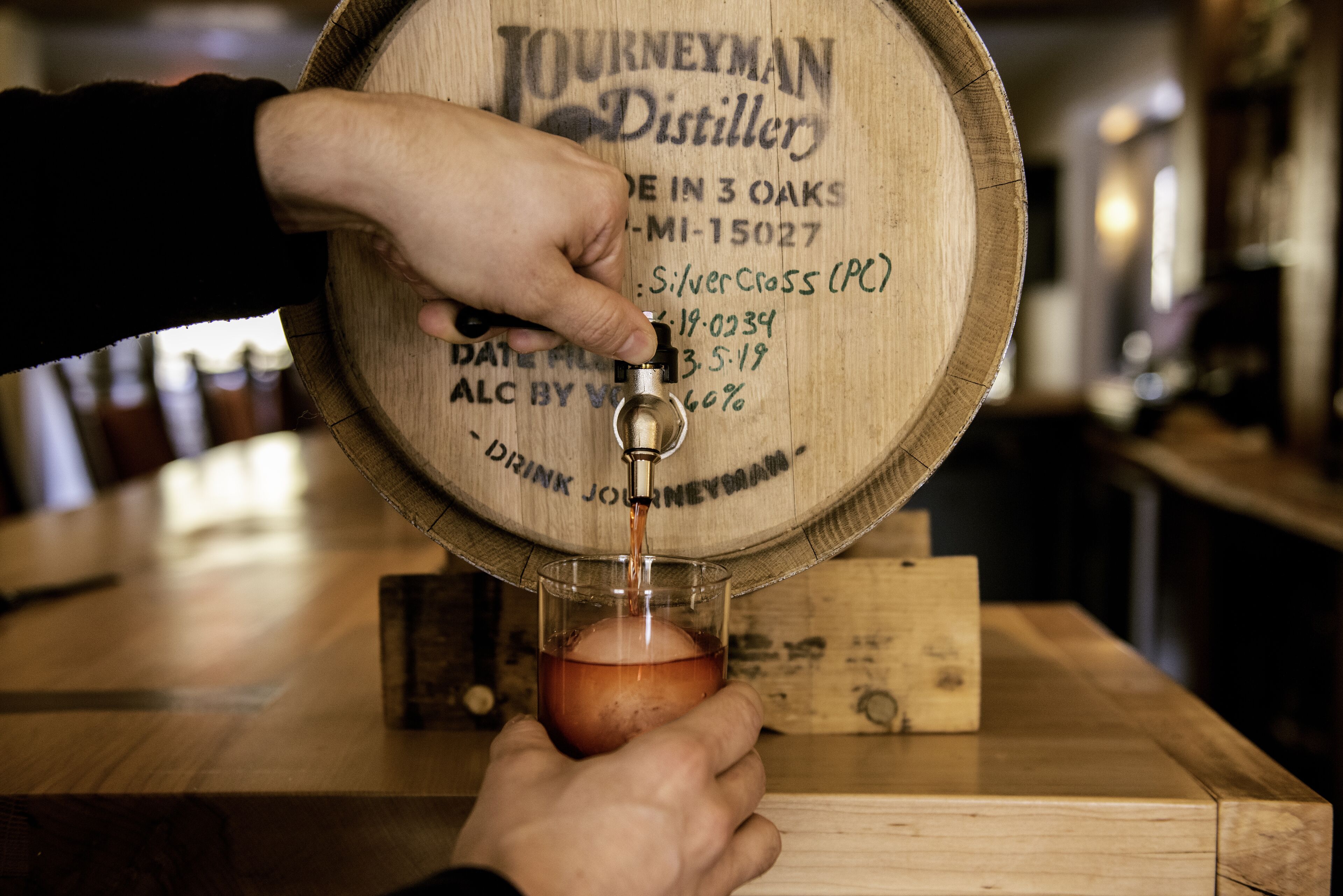 A hand pouring beer from a wooden barrel tap labeled "Journeyman Distiller" into a glass.