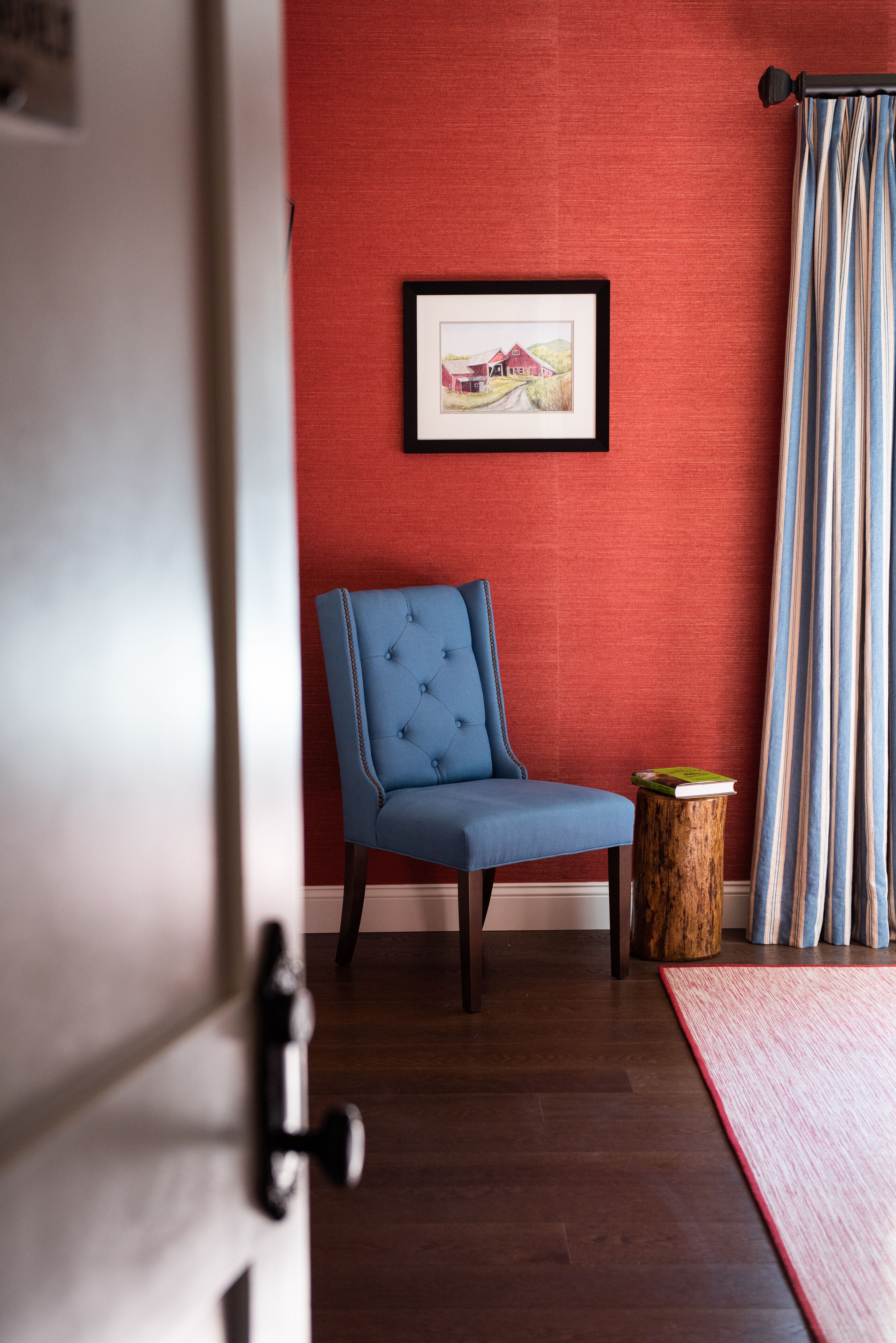 Ein Zimmer mit roter Wand, einem gerahmten Bild, einem blauen Stuhl, einem hölzernen Hocker und einem gestreiften Vorhang.