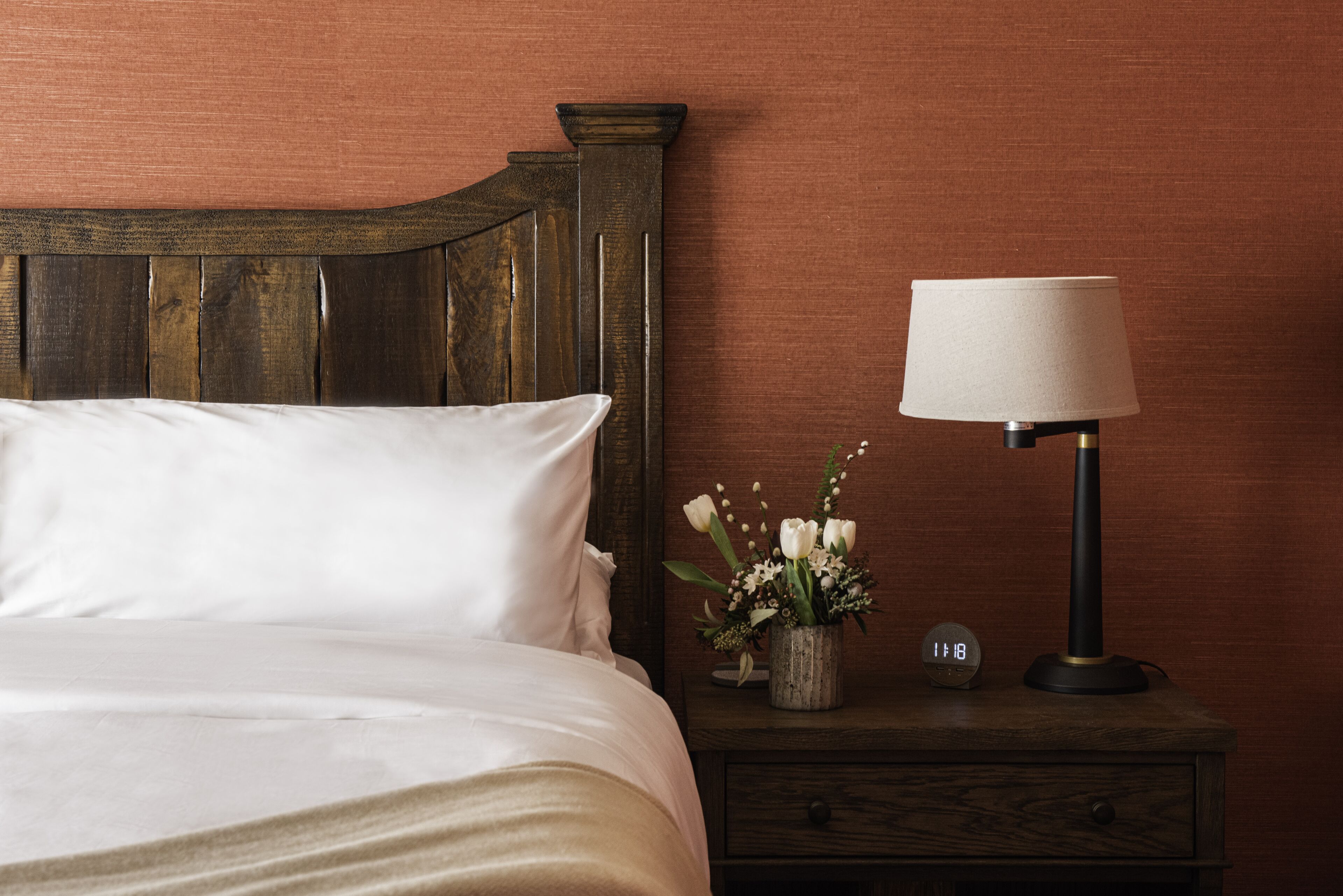 A neatly made bed with white linens, a wooden headboard, a bedside table with a lamp, a small clock, and a vase with flowers.