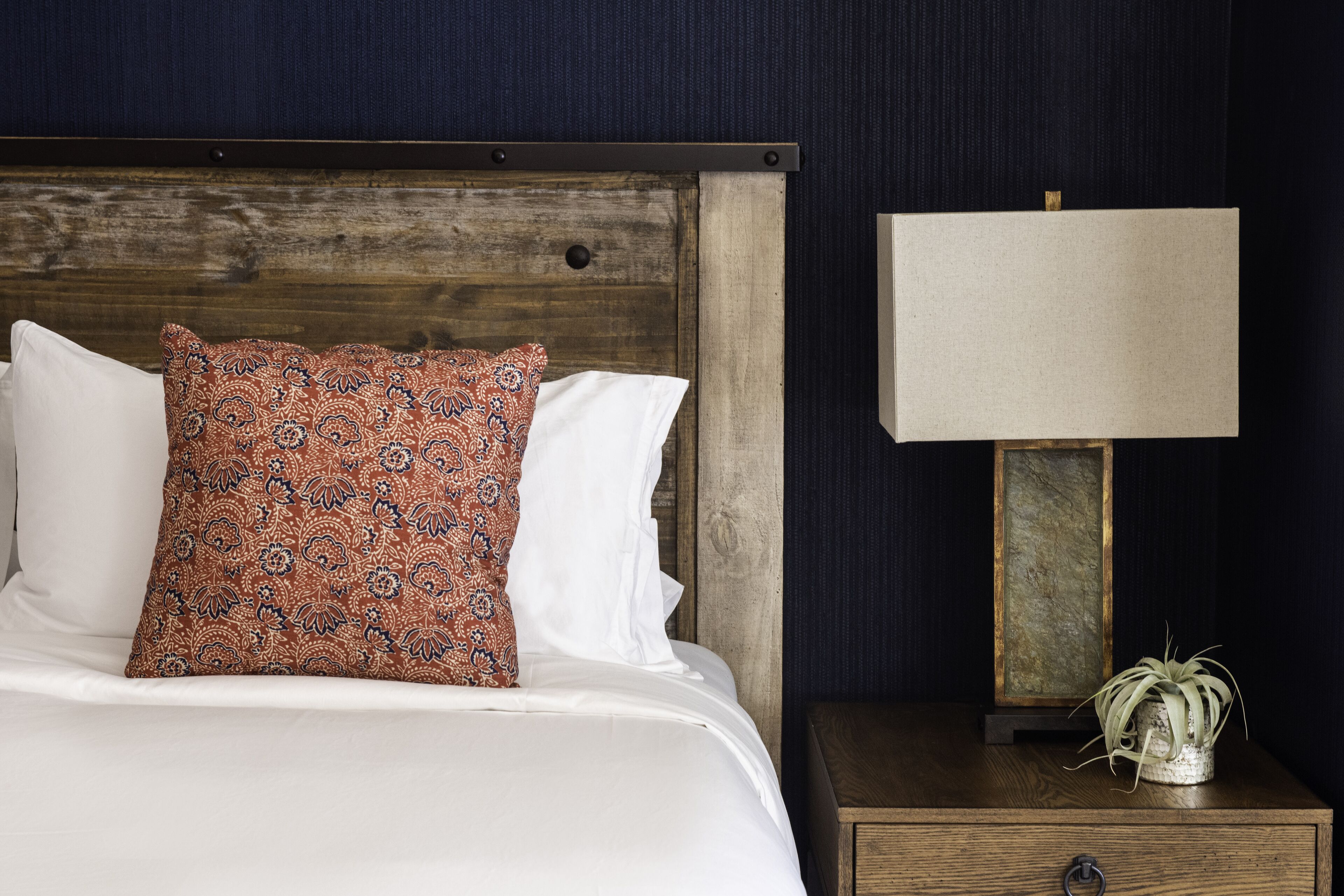 A neatly made bed with patterned and plain pillows, a wooden nightstand with a lamp and a small plant, and a dark blue wall.