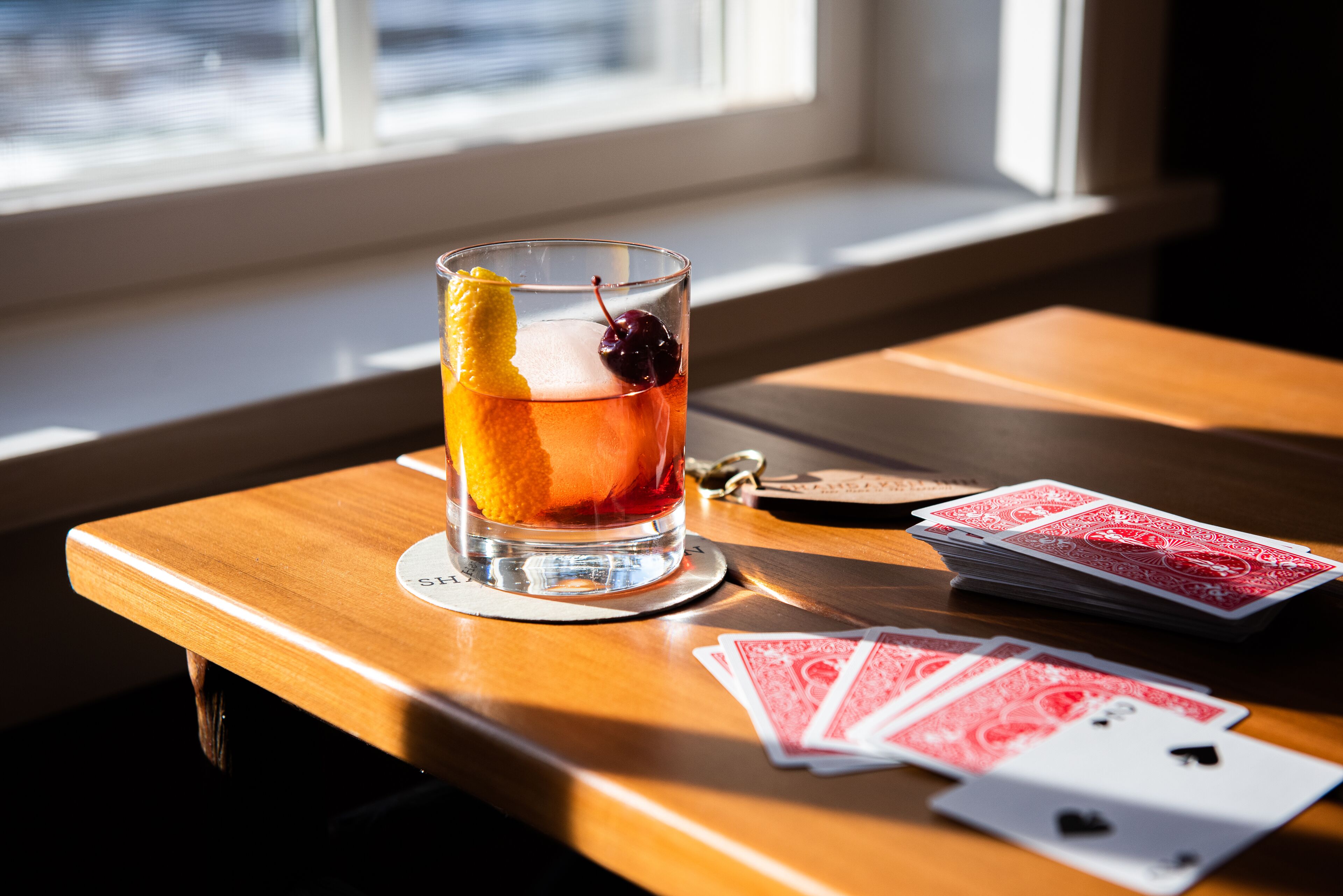 A glass of amber-colored drink with a cherry and orange peel garnish on a wooden table next to playing cards.
