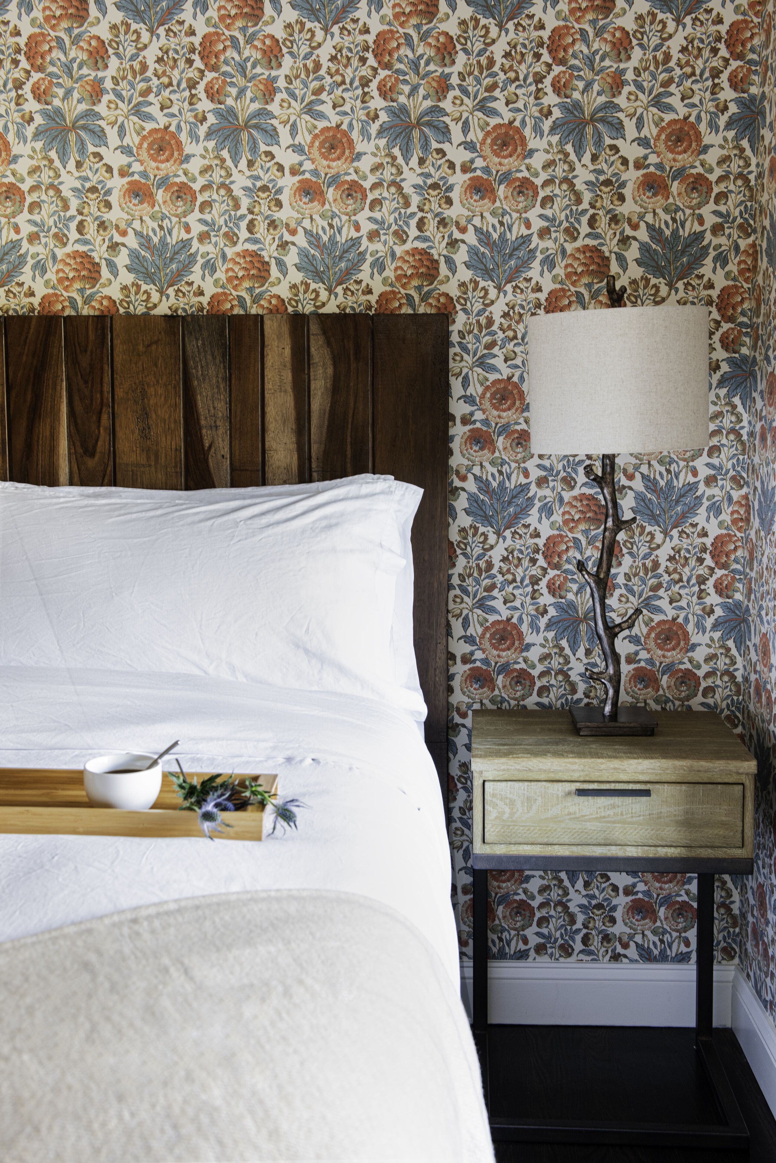Quarto com cabeceira de madeira, colcha branca, mesa de cabeceira com abajur e papel de parede estampado com flores.