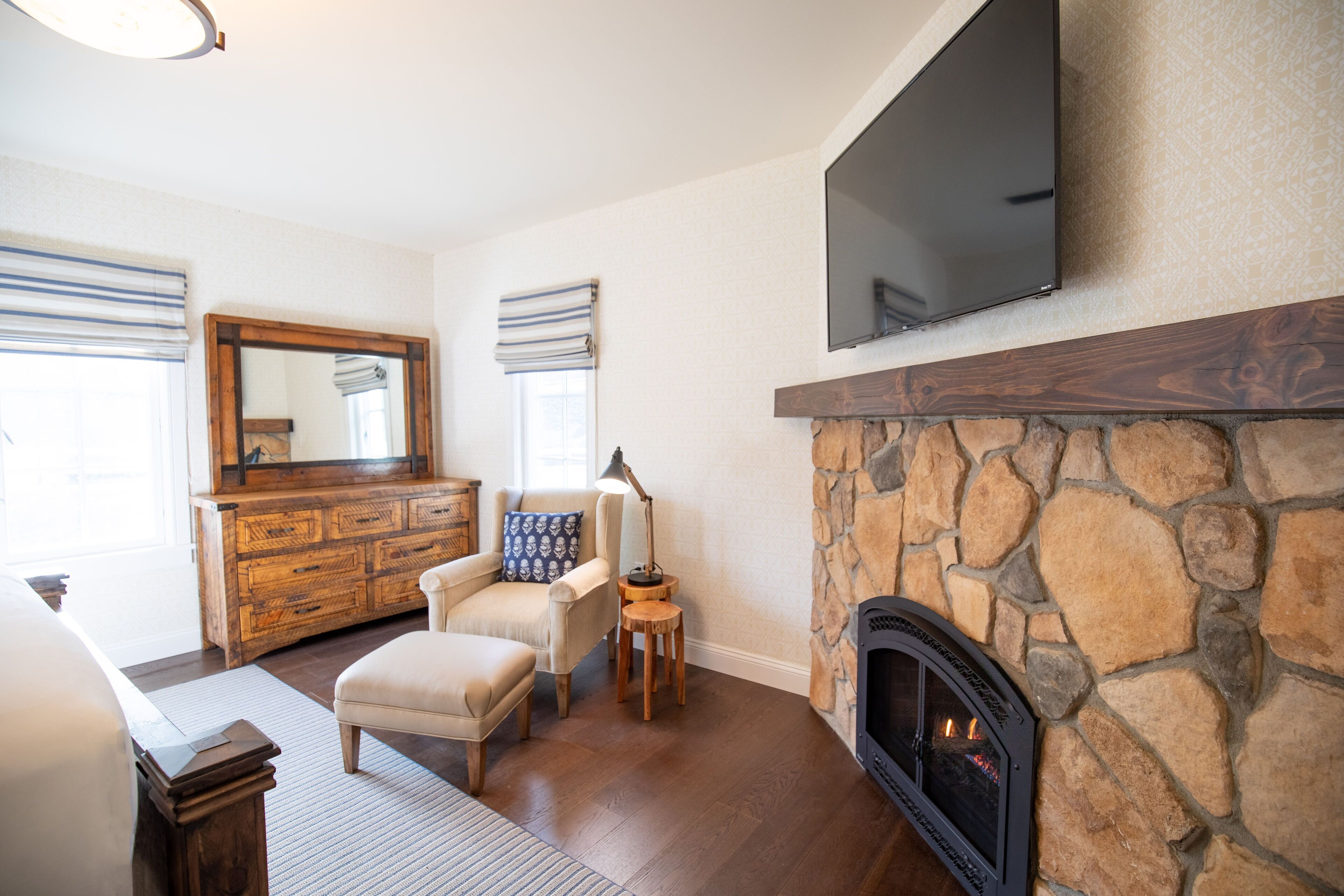 A living room with a stone fireplace, a television mounted above it, a wooden cabinet, and a comfortable seating area with a lamp.