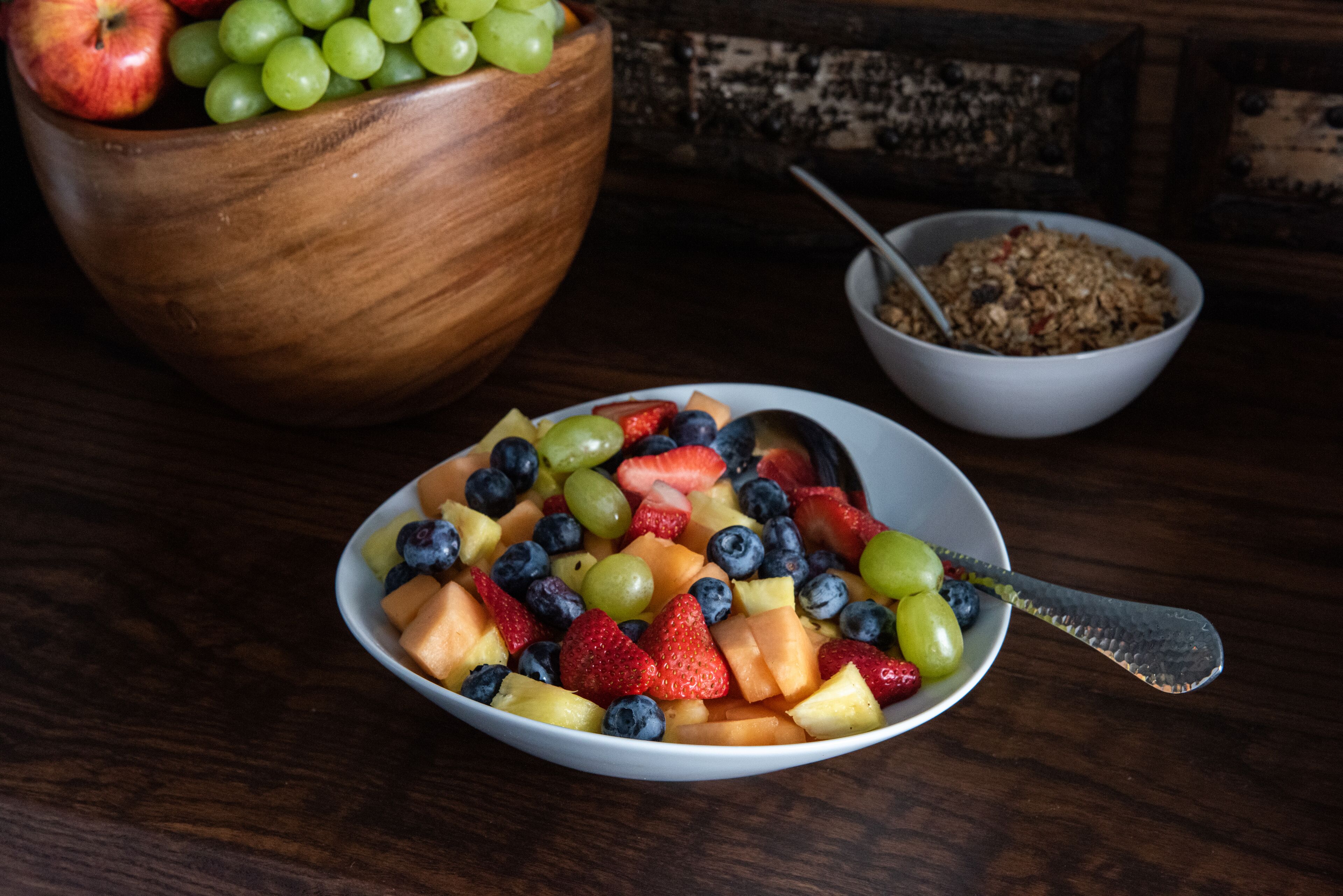 Een schaal fruitsalade met een lepel, een houten schaal met druiven, en een schaal granola op de achtergrond.
