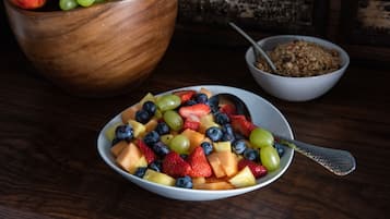 Een schaal fruitsalade met een lepel, een houten schaal met druiven, en een schaal granola op de achtergrond.