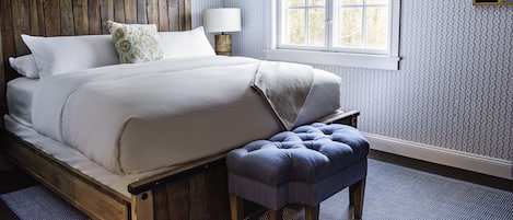 A bedroom with a wooden bed frame, white bedding, a blue upholstered bench, a bedside table with a lamp, and a window with a blue and white valance.