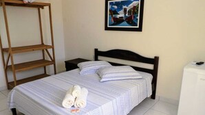 Una cama bien hecha con una colcha estampada y almohadas, un estante de madera, un cuadro en la pared y un electrodoméstico blanco a la derecha.