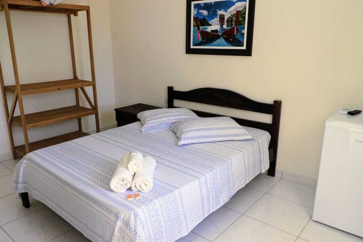 Un lit bien fait, avec une couvre-lit à motifs et des oreillers, une étagère en bois, un tableau encadré au mur et un appareil électroménager blanc sur la droite.