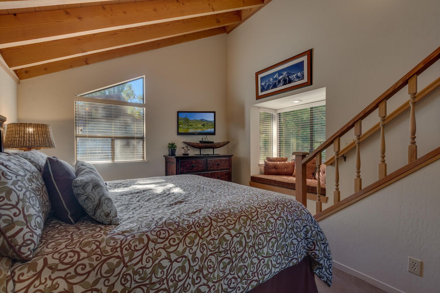 A bedroom with a large bed, a wooden ceiling, a TV, a desk, and a window with blinds.