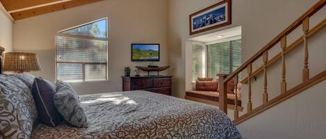 A bedroom with a large bed, a wooden ceiling, a TV, a desk, and a window with blinds.