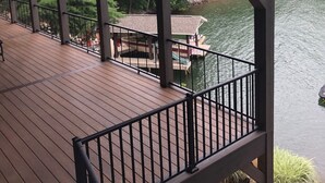 A wooden deck with a railing overlooking a body of water, a dock, and a small building with a roof.