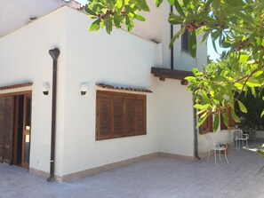 Un bâtiment à deux étages, doté d’une façade blanche, de volets en bois et d’une terrasse carrelée avec deux chaises en métal.