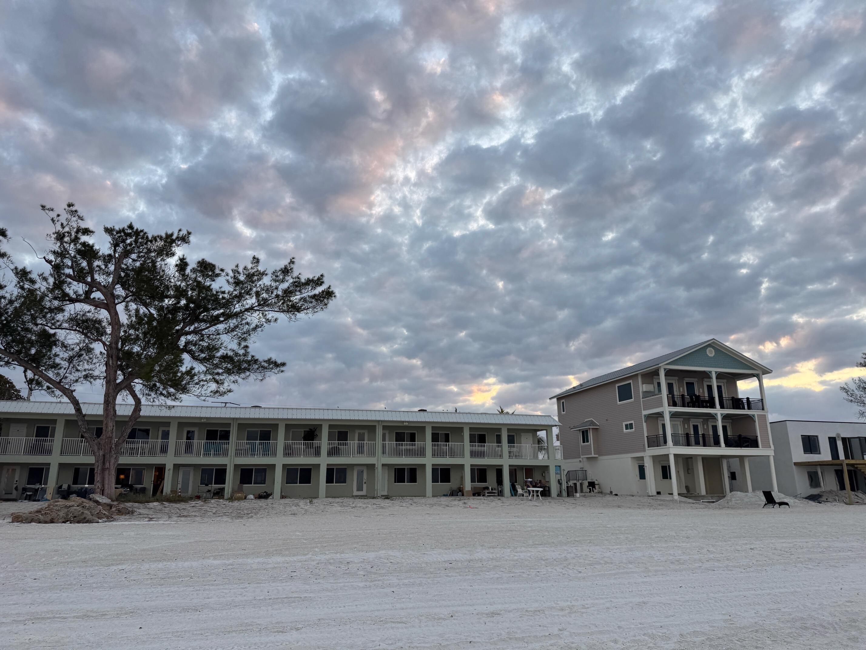 A multi-story building with a flat roof, a large open sandy area in front, and a tree to the left.