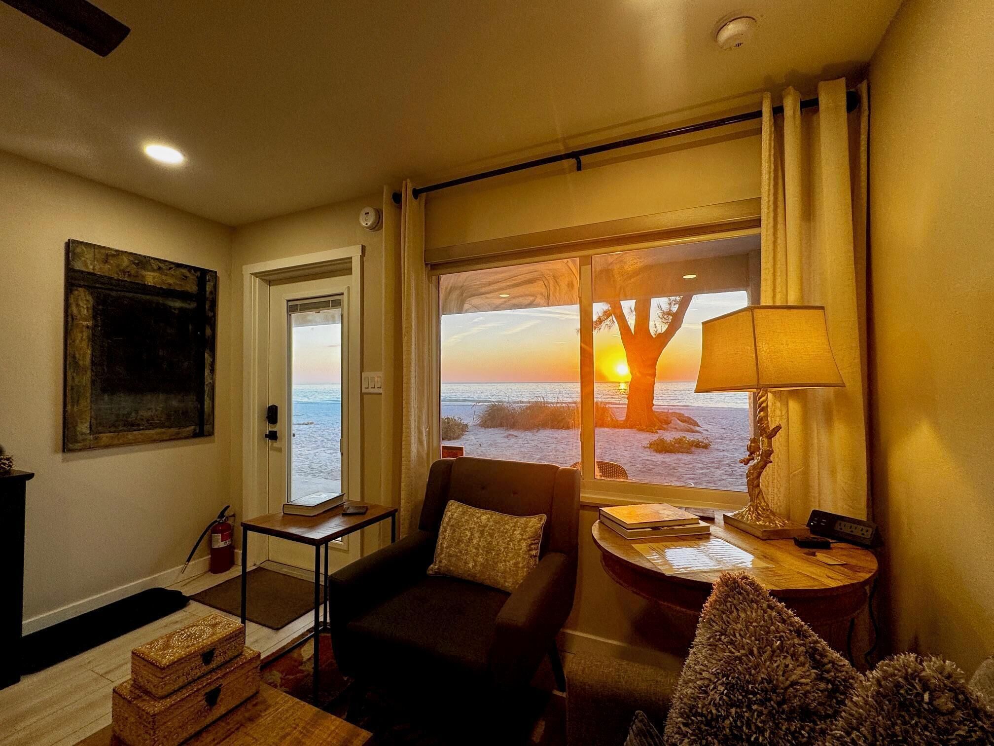 A cozy room with a view of the sunset over the ocean, a sofa, a small table with a lamp, and a fire extinguisher on the wall.