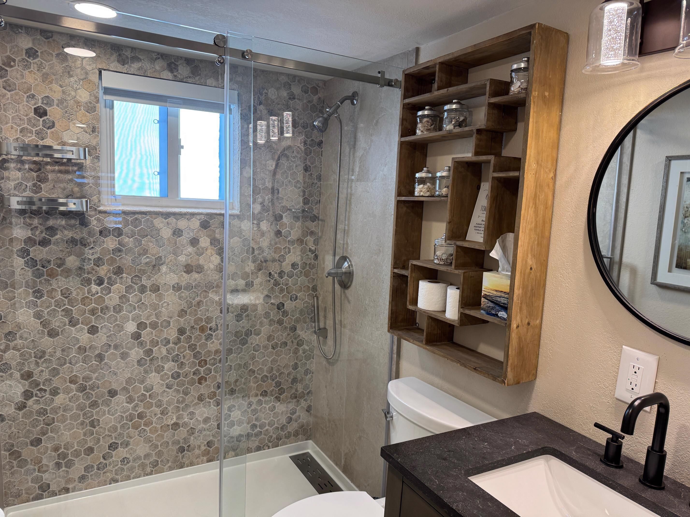 A modern bathroom with a glass-enclosed shower, a wooden shelf with toiletries, and a large mirror.
